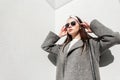 Glamorous young woman in stylish spring clothes straightens hair near wall on sunny day. Fine girl in trendy wear with sunglasses Royalty Free Stock Photo