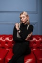 Glamorous young woman in trendy black clothes with a stylish pearl clips hair in a vintage pearl top is sits on a leather red sofa Royalty Free Stock Photo