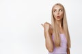 Glamour blond girl in tank top, pointing finger left and looking behind her shoulder with surprised face expression Royalty Free Stock Photo