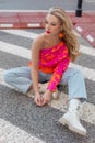 Glamour blonde with vivid make-up, in white boots, blue jeans and pink blouse sit on zebra crosswalk and look away. Royalty Free Stock Photo