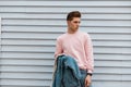 Glamour fashionable young man in pink stylish sweatshirt with denim blue jacket in jeans stands near white vintage building on Royalty Free Stock Photo