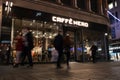 Exterior Night Shot of Caffe Nero Coffee Shop showing illuminated sign, logo and branding.  Blurred people walking past Royalty Free Stock Photo