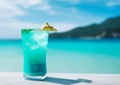 Glass with blue hawaiian cocktail on beach near crystal clear turquoise water against vibrant blue sky. Summer seaside vacation