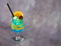 A glass of blue lagoon cocktail with a slice and a zest of orange, cherries, ice and a straw is on a gray background