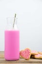Glass bottle filled with pink milk and a golden metal straw next to heart shaped cookies with pink frosting and sprinkles on