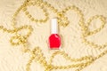 A glass bottle with red nail polish is on the yellow sand, golden beads are scattered randomly