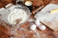 A glass bowl with flour and scapula on a wooden table sprinkled with flour. There are also other products on the table.