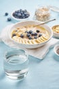 Glass of clear water and healthy diet breakfast with oatmeal, blueberries, banana on blue light background. Vertical. Side view