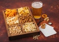 Glass of craft lager beer and opener with box of snacks on brown background. Pretzel,salty potato sticks, peanuts, onion rings