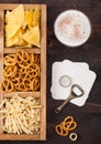 Glass of craft lager beer and opener with box of snacks on wooden background. Pretzel,salty potato sticks, peanuts, onion rings Royalty Free Stock Photo