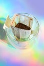 Glass cup with drip coffee bag on holographic rainbow background. Instant paper macro sachet above. Fresh morning making