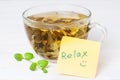 Glass cup with hot herbal tea and yellow sticker word relax on it mint leaves near in white wooden background macro