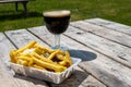 Glass of dark strong belgian beer and portion of fried potato chips with sauce and onion, served on outdoor terrace with green Royalty Free Stock Photo