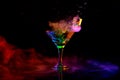 Glass with delicious cocktail with martini against dark background with neon light with smoke. Pouring liquid, splashes