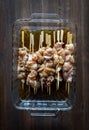 A glass dish of raw chicken marinading on bamboo skewers.