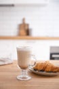 Glass of fragrant cappuccino coffee with cinnamon and croissant on a wooden table against the background of a white kitchen in th