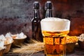 Glass with freshly poured beer and large head of foam near bottles, wheat and plates with snacks on dark wooden desk Royalty Free Stock Photo
