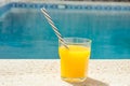 Glass of freshly pressed tropical fruits orange juice with striped straw standing on deck of swimming pool. Bright sunlight