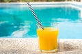 Glass of freshly pressed tropical fruits orange juice with striped straw standing on deck of swimming pool. Bright sunlight