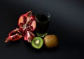 A glass of freshly squeezed fruit juice on a black background, next to a ripe kiwi fruit and a broken pomegranate fruit with seeds Royalty Free Stock Photo