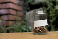Glass jar with coins inside and with a label with a wooden background, plants and bricks. Royalty Free Stock Photo