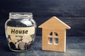 A glass jar with coins and a wooden house and the inscription `House`. The accumulation of money to buy a house. Rent an apartment