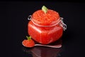 A glass jar filled with red caviar with a spoon with red caviar next to a dark, reflective background. Caviar, Russian food