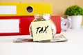 A glass jar full of coins sits on a stack of dollar bills on a work table. There is a sticker with the text TAX. Royalty Free Stock Photo
