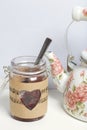 Glass jar of jam wrapped in paper with notes. Heart cut out in paper. Near a white kettle with roses Royalty Free Stock Photo