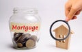 A glass jar with money and the word Mortgage and a wooden house. The accumulation of money to pay interest rates on mortgages.