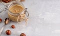 Glass jar with natural nut butter, spoon, healthy cookies, hazelnuts on a light background. Vegetarian traditional breakfast close