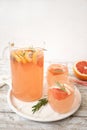 A glass jug and two glasses full of lemonade with grapefruit slices and sprigs of rosemary stand on a tray on a light wooden table