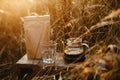 Glass kettle with coffee, cup and brown pack with coffee on background of rural countryside herbs in sunset. Alternative coffee