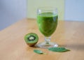 Glass of kiwi smoothie, half of a kiwi and green leaves on a wooden table with blur background