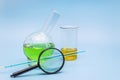 Glass laboratory flask and beaker in a cup with a green and yellow solution with a magnifying glass on a gentle blue medical Royalty Free Stock Photo