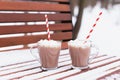 Glass mugs, cups with hot chocolate, cocoa, marshmallows. Winter snow covered park bench. Striped red, white paper straws for Royalty Free Stock Photo