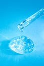 Glass pipette with facial serum, bubbles and falling drop on blue background closeup. Transparent dropper with liquid