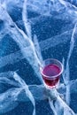 A glass with red liquor stands on the ice of a lake with beautiful cracks.