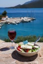 A glass of red wine and bowl of greek salad with greek flag on by the sea view, summer greek holidays concept. Royalty Free Stock Photo