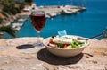 A glass of red wine and bowl of greek salad with greek flag on by the sea view, summer greek holidays concept.