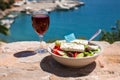 A glass of red wine and bowl of greek salad with greek flag on by the sea view, summer greek holidays concept. Royalty Free Stock Photo