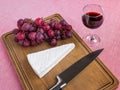 Glass of red wine and brown wooden cutting board with a triangular piece of Brie cheese, red sweet grapes, chef knife on it. Soft