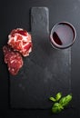 Glass of red wine, meat appetizer and basil on black slate stone board over dark background