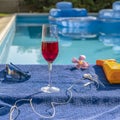 A goblet of red wine by the pool on a blue towel with sunglasses, earphones, a pink oleander flower and sunscreen next to it Royalty Free Stock Photo