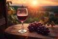 A glass of red wine and purple grapes on a wooden table with a view of the vineyard at sunset. Royalty Free Stock Photo