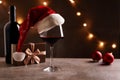 Glass of red wine with Santa Claus hat, bronze gift box and Christmas decorations on table. Christmas lights in the black