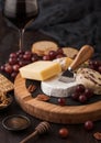 Glass of red wine with selection of various cheese on the round board and grapes on wooden background. Blue Stilton, Red Leicester