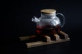 Glass teapot with hot black tea on a wooden stand for hot dishes. Perspiration on the glass walls of the teapot. Black background