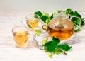 A glass teapot and two cups of tea with a linden tree on a wooden table against a background of linden leaves and Royalty Free Stock Photo