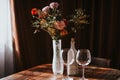 Glass vase with flowers on a wooden table in front of a light background. On the table are a bottle and glass wine glasses. Royalty Free Stock Photo